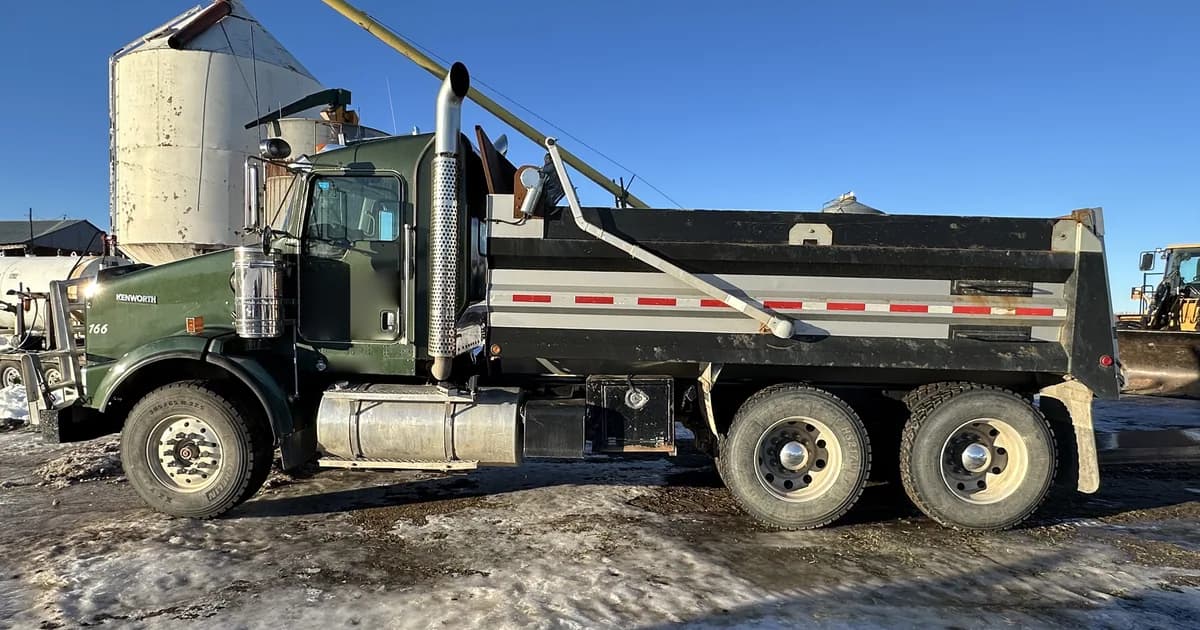 A green 2011 Kenworth T800 tandem-axle dump truck in side profile on a snowy Alberta farm yard, beside a grain bin