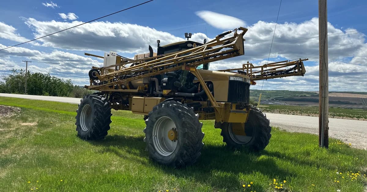 A 2007 RoGator 1074SS high-clearance sprayer with booms folded, parked on grass beside a cultivated prairie field under a big Canadian sky