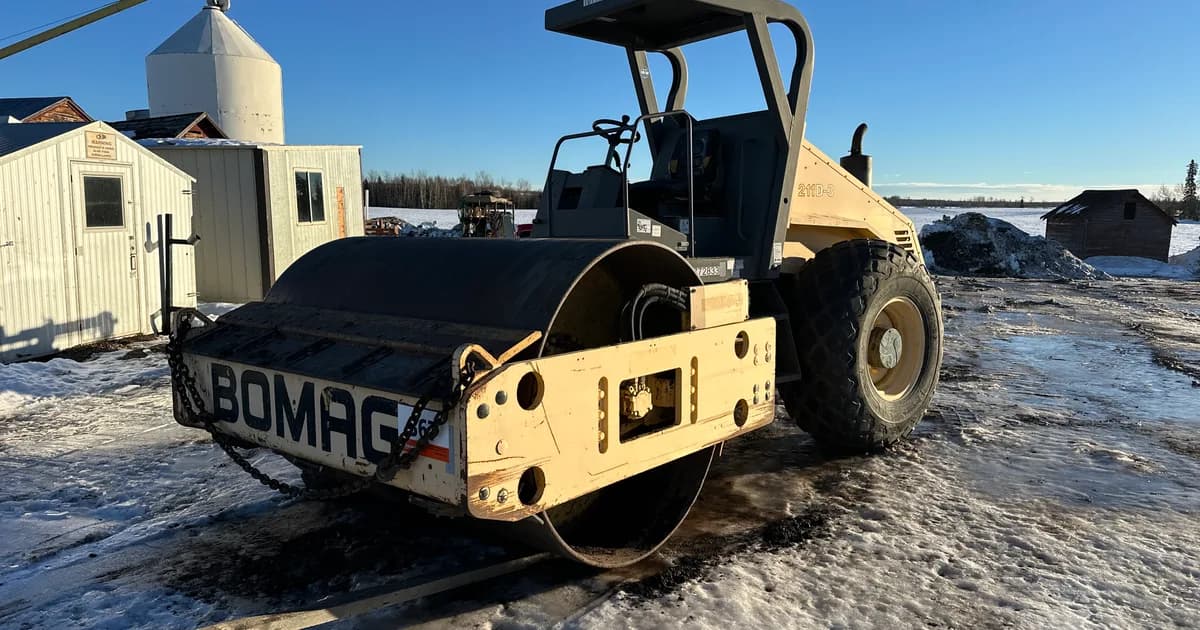 A Bomag smooth-drum compactor parked in a snowy Alberta farm yard beside grain bins — an owner-held machine of the kind bought and sold privately between contractors